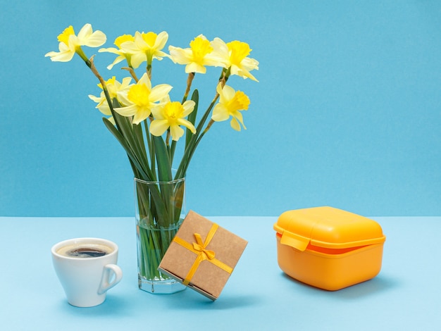 Bouquet di narcisi gialli in vaso di vetro, una confezione regalo, una tazza di caffè e una scatola per il pranzo su superficie blu blue