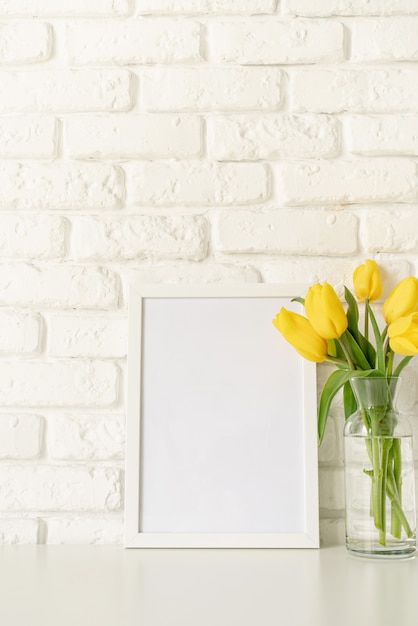 Bouquet di tulipani gialli in un vaso di vetro e una cornice vuota su uno sfondo di muro di mattoni bianchi. Mock up design