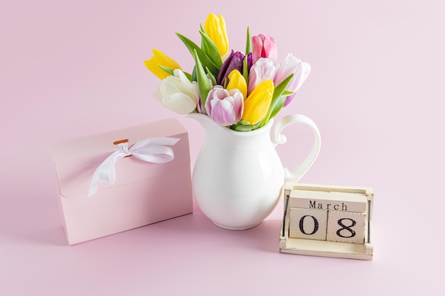 Bouquet di tulipani primaverili in una brocca bianca su uno sfondo rosa accanto a una scatola di vacanza e un calendario in legno con la data dell'ottava festa di marzo