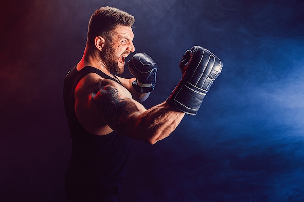 Boxer muay thai sportivo tatuato barbuto in canottiera nera e guantoni da boxe combattendo sul muro scuro con fumo. Concetto di sport.