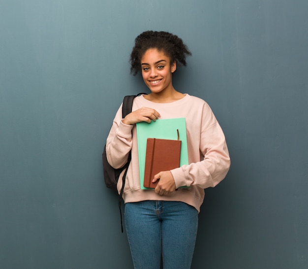 Braccia dell'incrocio della giovane donna di colore dello studente, sorridenti e rilassate. Tiene dei libri.