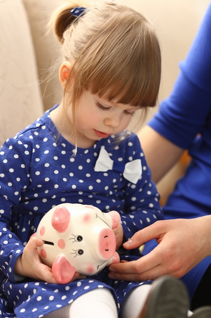 Braccio della bambina del bambino che mette le monete nel salvadanaio