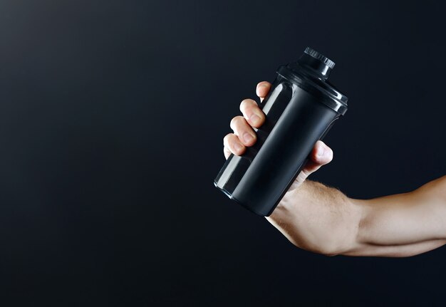 Braccio di uno sportivo muscoloso che allunga una mano in avanti con uno shaker nero con proteine su sfondo scuro.