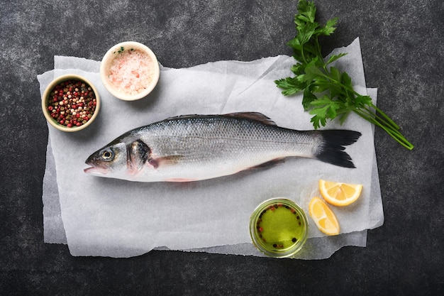 Branzino crudo Pesce di mare fresco Spigola con sale, pepe, prezzemolo, olio d'oliva e limone su carta pergamena su fondo rustico di cemento scuro Sfondo di cottura degli alimenti Vista dall'alto dello spazio della copia