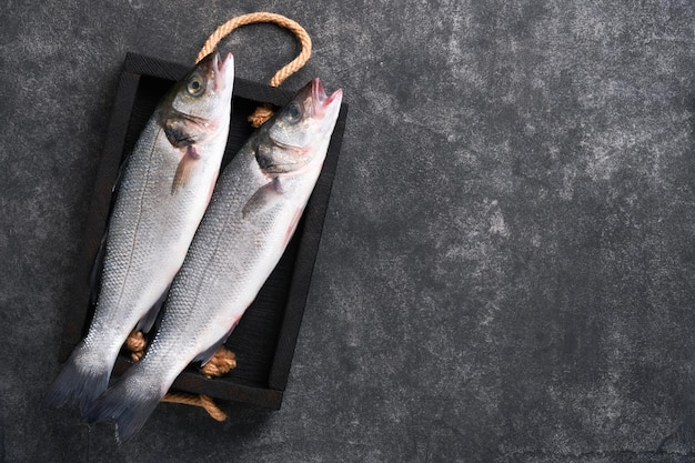 Branzino crudo Pesce di mare fresco Spigola con sale, pepe, prezzemolo, olio d'oliva e limone sul tagliere su fondo rustico in cemento scuro Sfondo di cottura degli alimenti Vista dall'alto dello spazio della copia