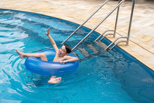 Brasiliano di 9 anni in piscina in una giornata di sole, con la mano alzata, seduto su una boa e incollato