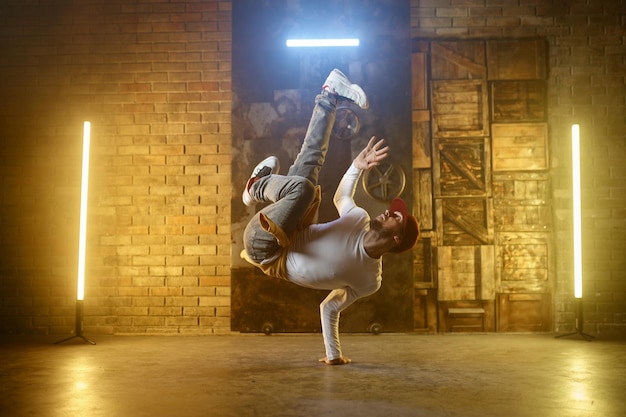 Breakdance del giovane che fa handstand con una mano. Ballerino hip-hop attivo ed elegante che si allena o si esibisce su sfondo di studio loft