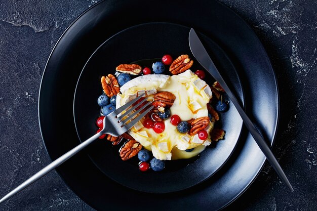 Brie al forno con noci pecan d'acero e frutti di bosco: mirtilli, mirtilli rossi e miele in cima serviti su un piatto nero