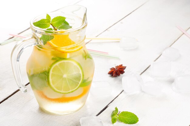 Brocca con lime e frutta arancione Acqua con ghiaccio e menta su tavola bianca