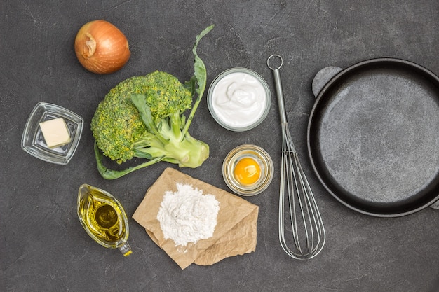 Broccoli e farina, uovo rotto in una ciotola di vetro, olio d'oliva, cipolla e frusta. Padella sul tavolo. Sfondo nero. Lay piatto