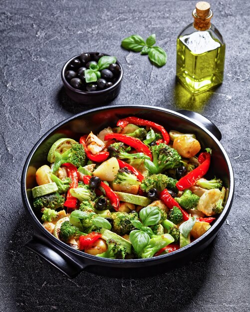 Broccoli e pollo al forno con patate novelle, peperoncino, zucchine, olive nere con foglie di basilico fresco sopra, servito su una teglia nera su fondo di cemento, vista dall'alto, primo piano, vista verticale