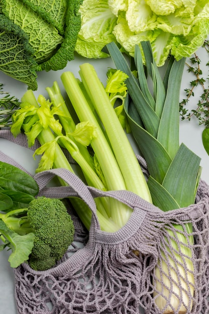 Broccoli, porri e gambi di sedano in un sacchetto di cordoncino. Cavolo cinese e verza sul tavolo. Disposizione piatta. Sfondo grigio