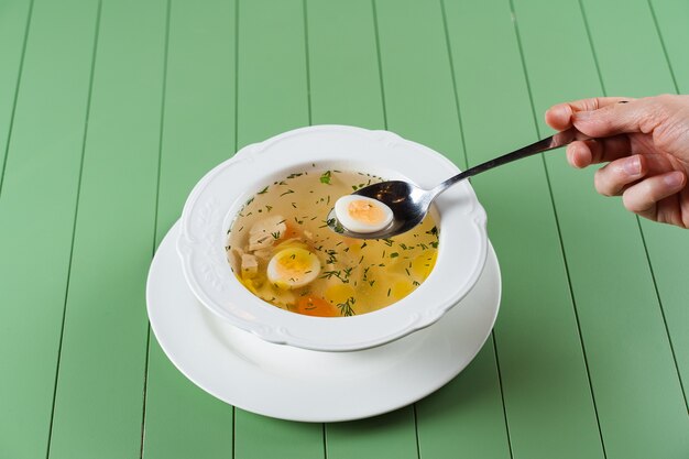 Brodo di pollo con carne, carote, erbe aromatiche e uovo di quaglia in un piatto bianco su un tavolo verde