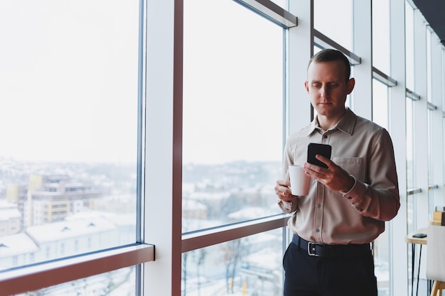 Broker maschi con il telefono che guarda verso la finestra con il caffè in mano nell'area di lavoro moderna