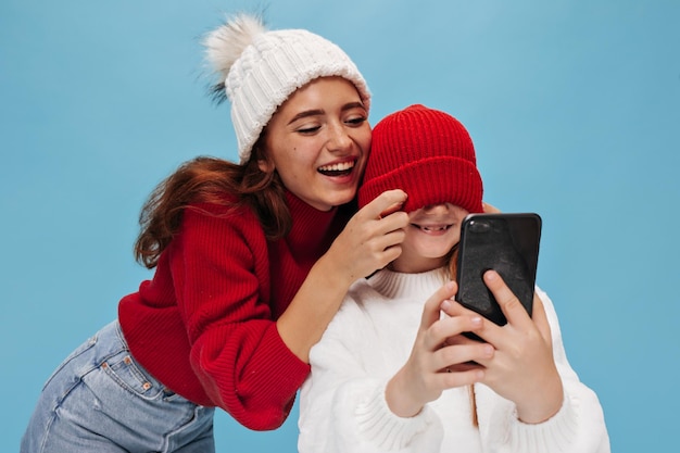 Bruna allegra con cappello bianco e maglione rosso ride e chiude gli occhi sorellina in abiti moderni su sfondo isolato