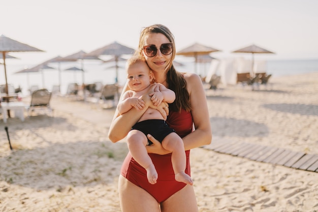 Bruna caucasica attraente felice vestita in costume da bagno che tiene il suo allegro figlio di 6 mesi mentre si trovava su una spiaggia.