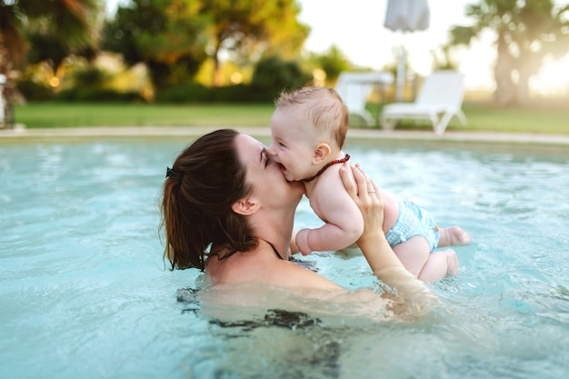 Bruna caucasica dedicata attraente che bacia il suo figlio amorevole di 6 mesi mentre levandosi in piedi in piscina. Prima volta al concetto di piscina.