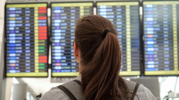Bruna con coda di cavallo e zaino grigio che indossa una t-shirt guarda l'orario delle partenze nella vista sul retro della lounge dell'aeroporto