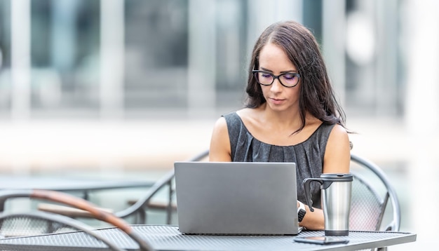 Bruna con gli occhiali lavora all'aperto circondato da edifici per uffici su un laptop