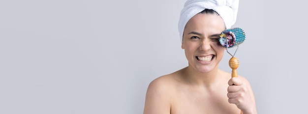 Bruna con un asciugamano in testa usando il rullo per il massaggio del viso isolato su sfondo grigio