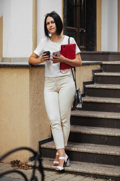 bruna donna d'affari si trova vicino all'edificio della città con tablet, telefono e bevande caffè