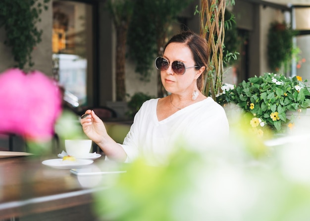 Bruna donna di mezza età in occhiali da sole in abiti bianchi con caffè sulla terrazza del caffè estivo