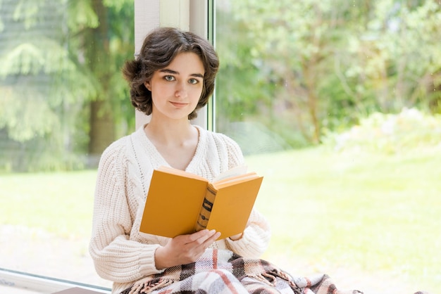 Bruna donna in maglione caldo libro di lettura seduto in camera sulla veranda