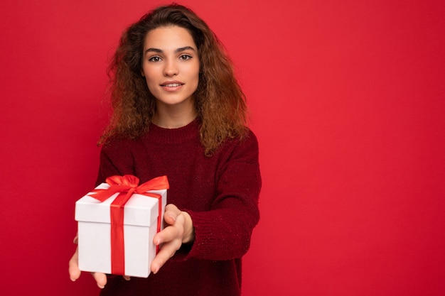 bruna donna riccia isolata su sfondo rosso muro che indossa un maglione rosso azienda confezione regalo guardando la fotocamera.