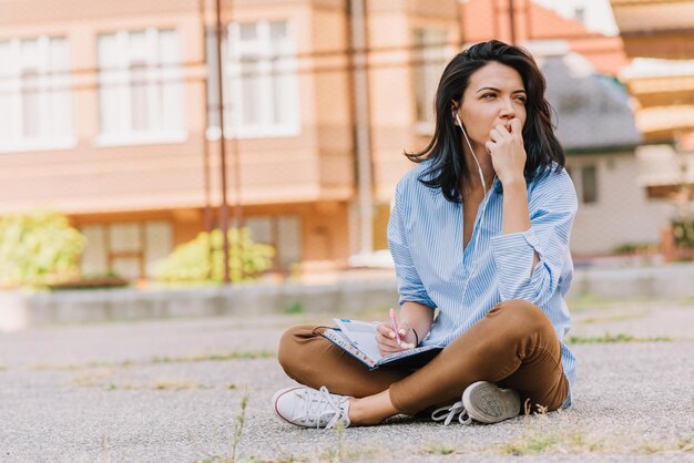 Bruna europea bella donna seduta sul marciapiede a studiare e prendere nota sul pianificatore di notebook Giovane donna carina con gli auricolari ascoltare musica e prepararsi per l'esame Concetto di persone e stile di vita