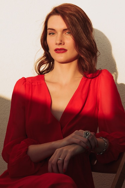 Bruna giovane ragazza in piedi sul balcone al tramonto in un vestito rosso e occhiali da sole