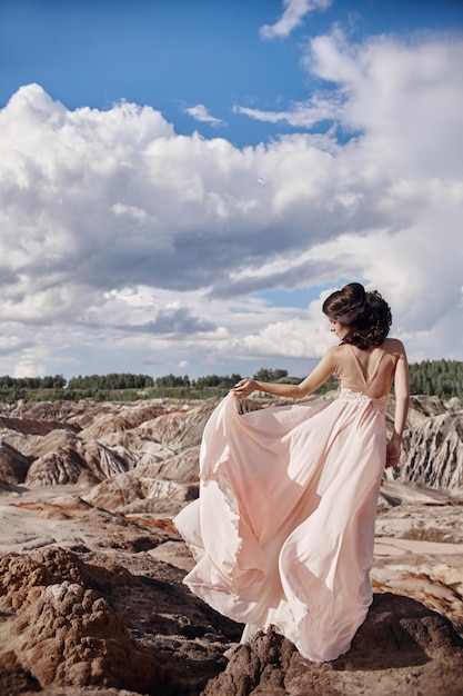 Bruna in abito rosa si erge su una scogliera, il vento scompiglia un vestito, una tenera immagine di una ragazza. Favoloso paesaggio di montagna. Donna nel vento