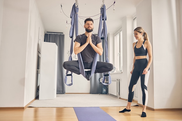 Bruna maschio caucasico che fa asana in amaca in studio mentre una donna bionda esile lo osserva