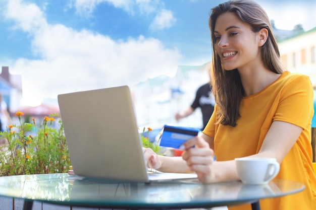 Bruna naturale felice che utilizza carta di credito e laptop nella caffetteria all'aperto