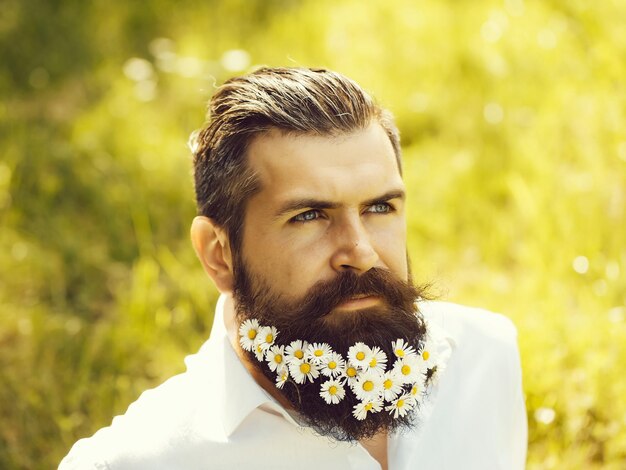 Bruna uomo bello hipster con fiori di margherita in barba in camicia nera con baffi eleganti e capelli alla moda giornata di sole all'aperto