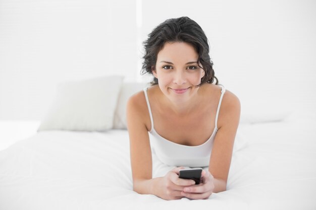 Brunette casuale allegro in pigiami bianchi che manda un sms con un telefono cellulare