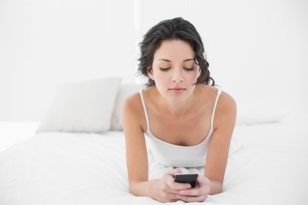 Brunette casuale rilassato in pigiami bianchi che texting con un telefono cellulare