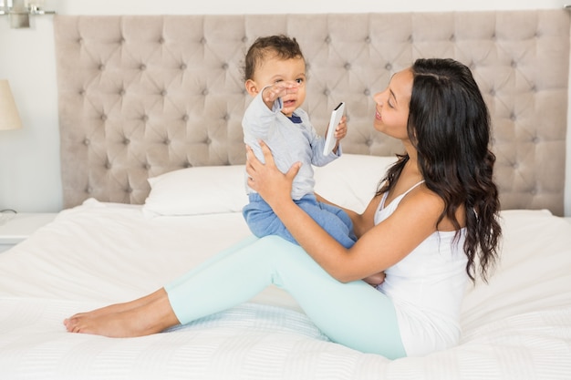 Brunette felice che tiene il suo bambino che sta tenendo smartphone sul letto