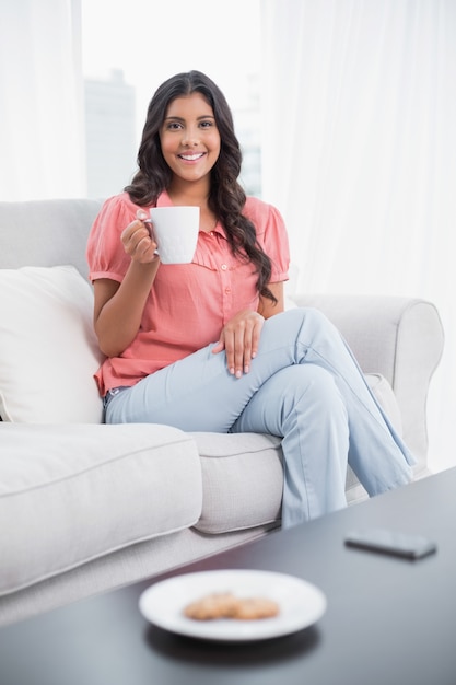 Brunette sveglio sorridente che si siede sulla tazza della holding dello strato