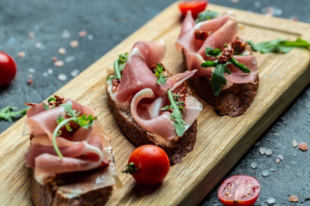 Bruschetta con pomodori secchi, prosciutto e rucola su una tavola di legno. Deliziosa colazione o merenda. Sfondo di ricetta alimentare. Avvicinamento.