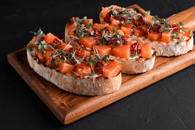 Bruschetta con salmone, pomodori secchi, erbe aromatiche, ricotta, su fondo nero, tavola di legno