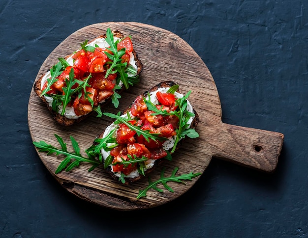 Bruschetta di pane integrale di pomodori e rucola su un tagliere di legno su fondo scuro