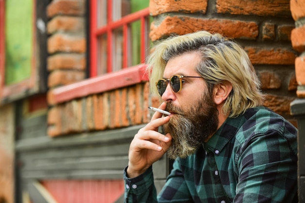 Brutale maschio caucasico con gli occhiali uomo che fuma all'aperto alla moda uomo maturo che fuma sigaretta punk hipster uomo che fuma fumo e abitudine fumo passivo o di seconda mano copia spazio