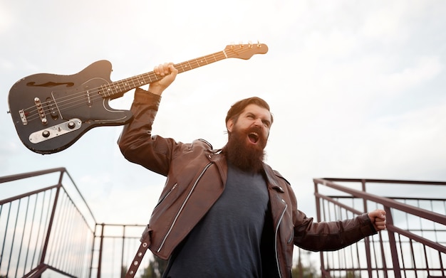 Brutale rocker barbuto con i capelli grigi in una giacca di pelle marrone e jeans blu tiene una chitarra elettrica in mano e grida