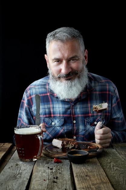 Brutale uomo adulto dai capelli grigi con la barba mangia bistecca di senape e beve birra