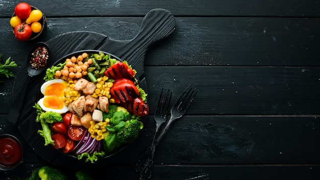 Buddha Bowl pollo ceci broccoli pomodori uovo paprika cipolla in un piatto nero su sfondo nero Vista dall'alto Spazio libero per il testo