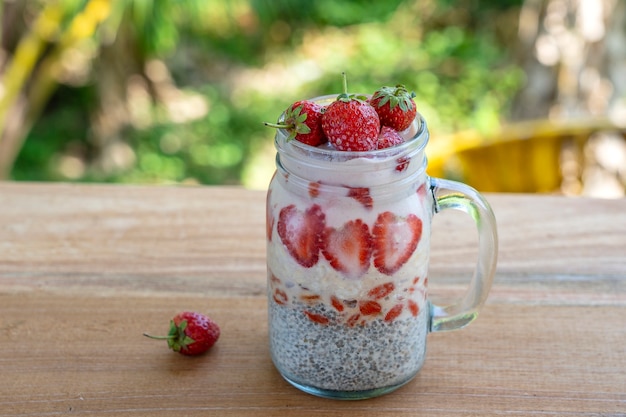 Budino di chia al latte di mandorle con fragole rosse fresche, bacche di goji e fiocchi d'avena in una tazza di vetro. Colazione cruda vegana. Semi di chia e frutta fresca tagliata e dessert di bacche, primo piano