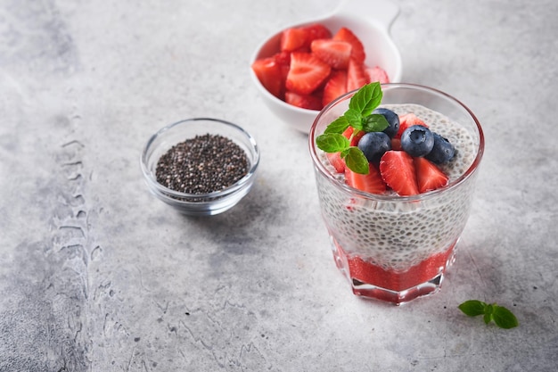 Budino di chia alla fragola Sana colazione vegana budino di semi di chia con frutti di bosco freschi e menta in vetro su sfondo grigio cemento Concetto di alimentazione sana dieta menu fitness Messa a fuoco selettiva