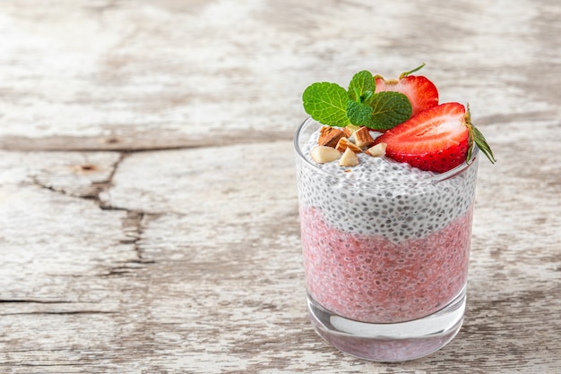 Budino sano di chia alla fragola e alla vaniglia in un bicchiere con frutti di bosco freschi, noci e menta su tavola rustica bianca