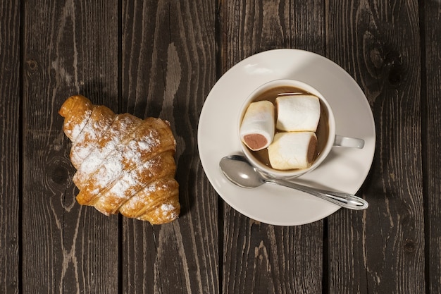 Bun croissant con caffè caldo con latte e gustosi marshmallow al cioccolato in una tazza bianca su uno sfondo di legno scuro. Lay piatto.