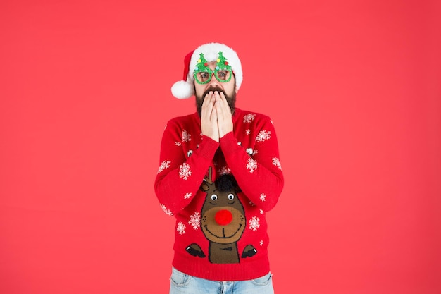 Buon anno. divertente maglione lavorato a maglia hipster. vestiti caldi per il freddo. stagione delle vacanze invernali. uomo barbuto santa cappello sfondo rosso. buon Natale. maschio sorpreso in occhiali da festa di Natale.